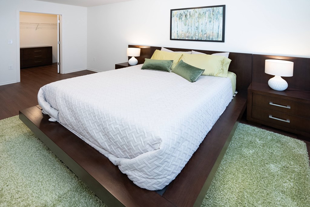 A bed with a white comforter and green pillows is in a room with a painting on the wall.