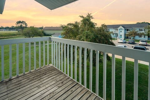A balcony with a wooden floor and white railings overlooks a parking lot and buildings.
