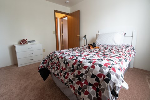 A bed with a white and red comforter in a bedroom with a white dresser and a brown carpet.
