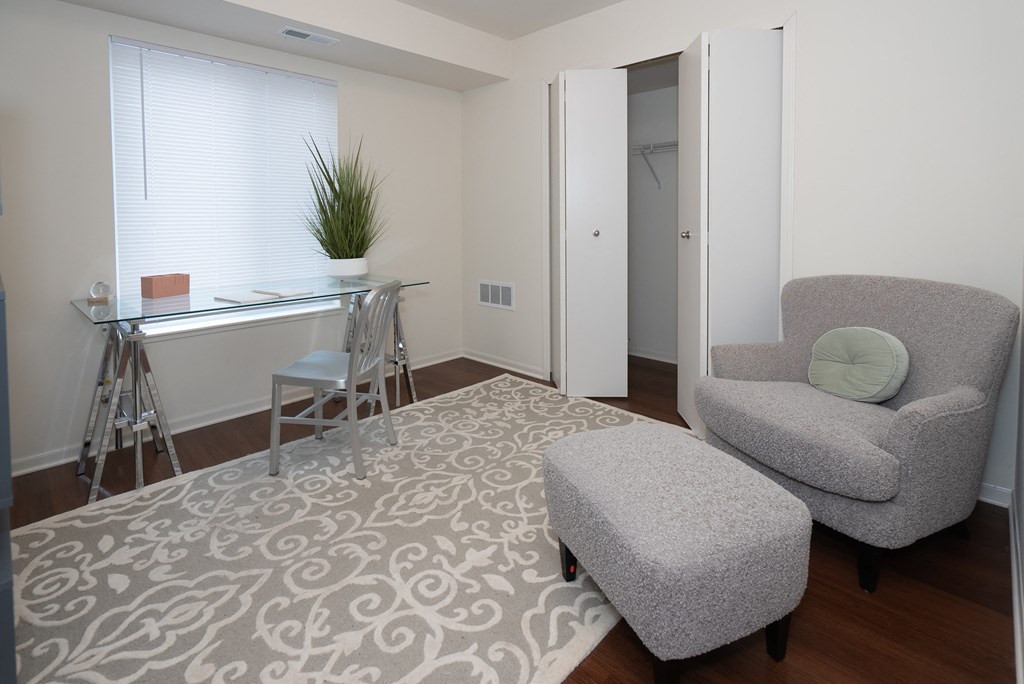A room with a grey chair and ottoman, a desk with a plant on it, and a patterned rug.