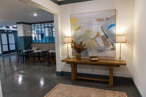 A large abstract painting hangs on the wall above a wooden bench with a vase of dried flowers on it.