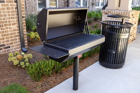 A black grill is placed on a sidewalk next to a trash can.