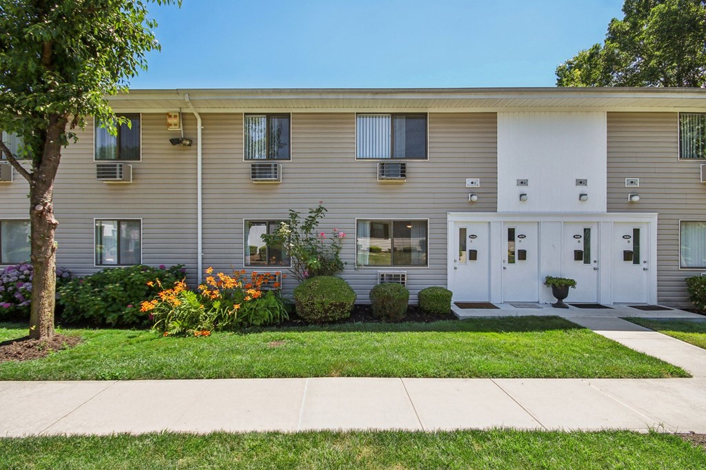 Exterior view of apartment building with green grass and trees at Brookwood at Ridge, New York, 11961