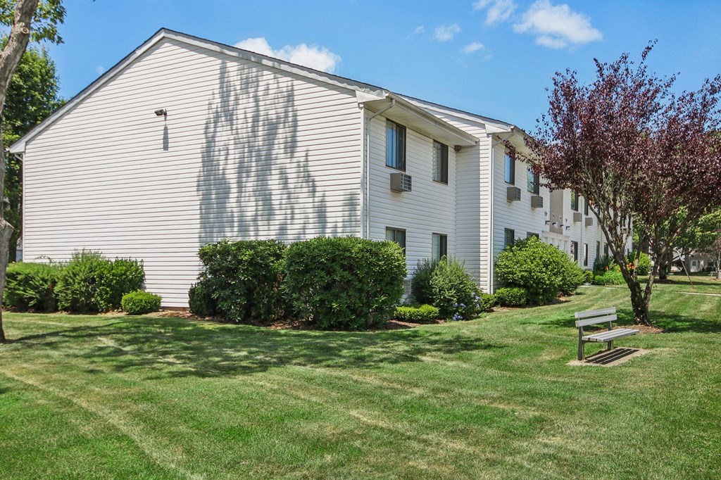 Exterior view of apartment building with green grass and bench seating at Brookwood at Ridge, New York, 11961