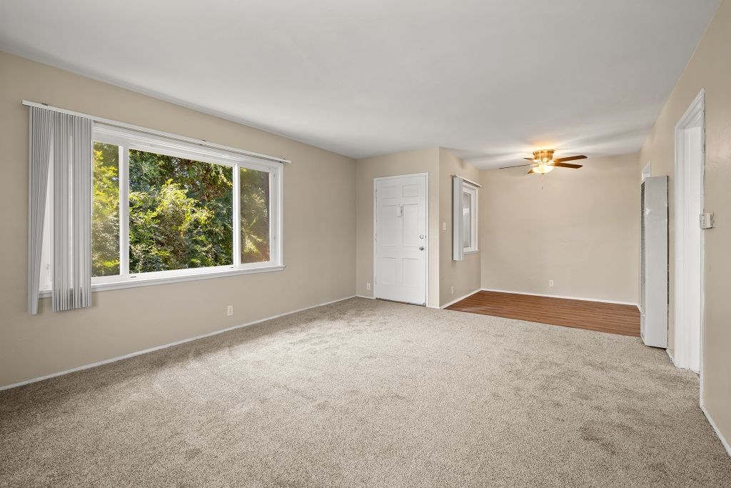 A room with a carpeted floor, a window with white curtains, and a ceiling fan.
