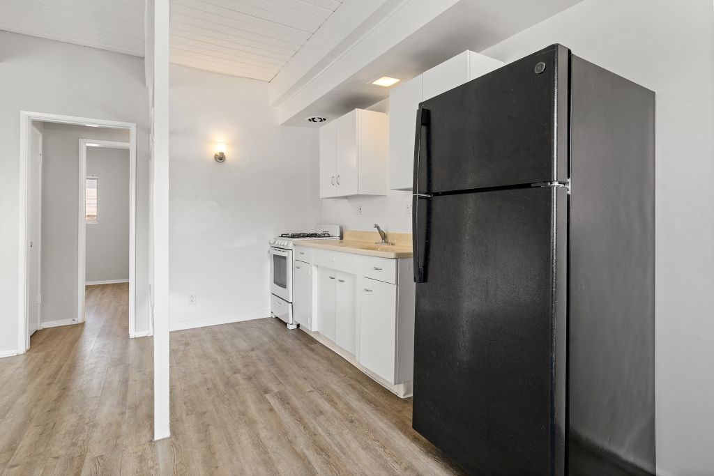 A black refrigerator stands in a kitchen with white cabinets and a wooden floor.