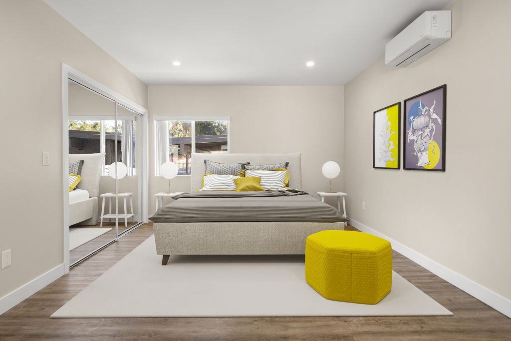 A bedroom with a bed, a yellow ottoman, and a painting on the wall.