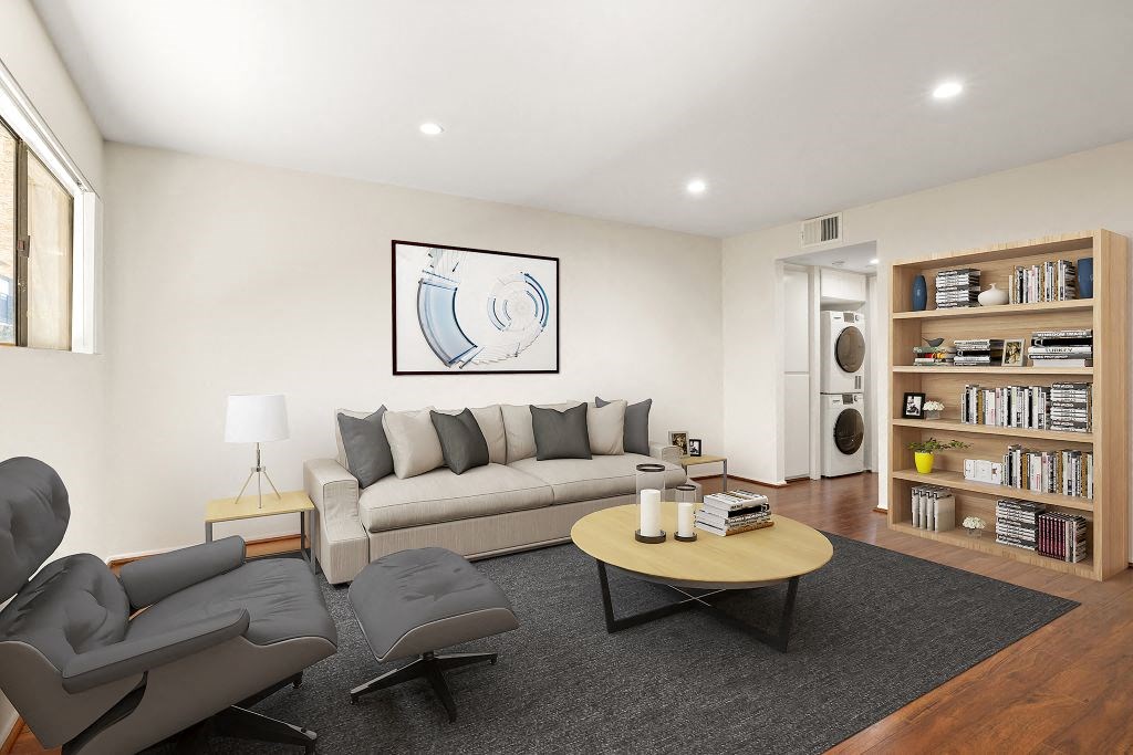 A living room with a grey couch, a grey chair, a black coffee table, a lamp, a book, a plant, a picture on the wall, a white door, and a bookshelf.