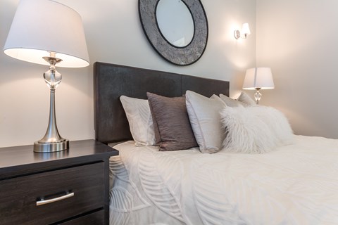A bedroom with a bed, nightstand with a lamp, and a round mirror on the wall.