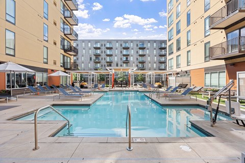 A large swimming pool surrounded by lounge chairs and umbrellas in a sunny courtyard.
