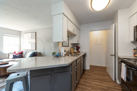A kitchen with a white countertop and grey cabinets, a living room with a window in the background
