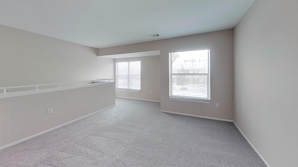 A spacious room with carpeted flooring and two large windows.