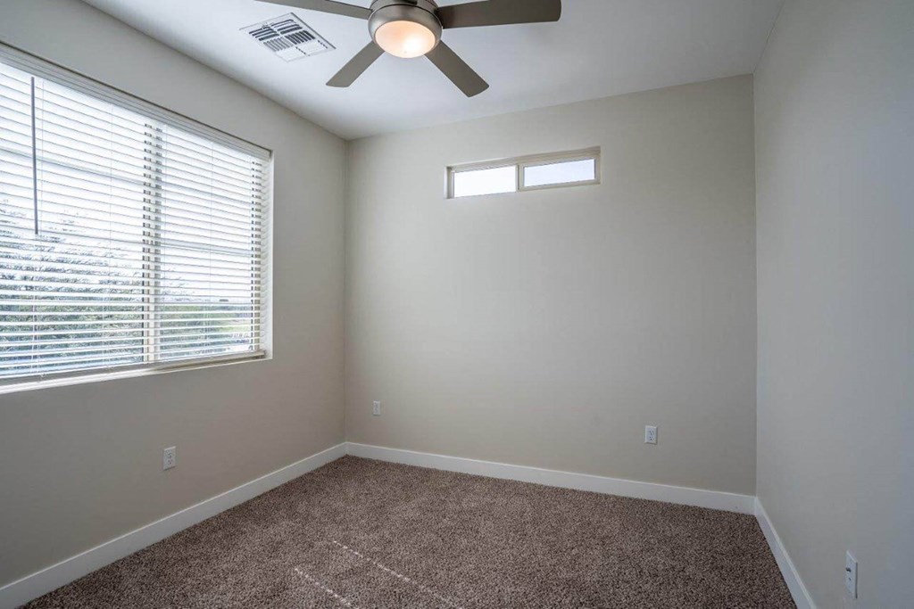 A carpeted bedroom has a window and ceiling fan in an apartment at Village Greens of Queen Creek