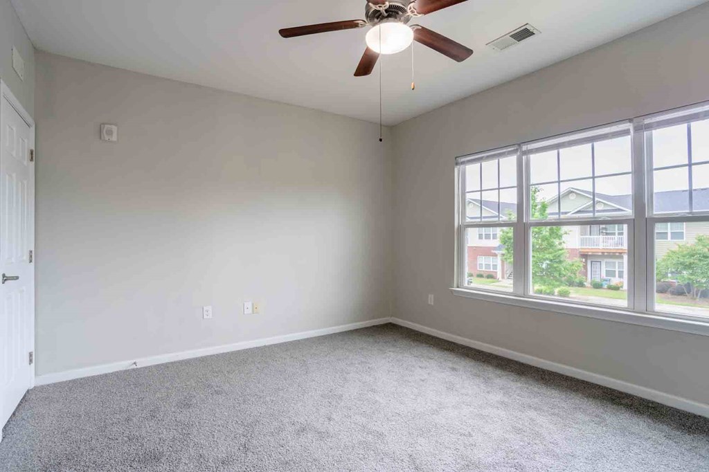 Large bedroom with neutral tones, carpet flooring, ceiling fan, and oversized windows offering abundant natural light at Cedar Terrace Apartments in Hendersonville, NC 28792