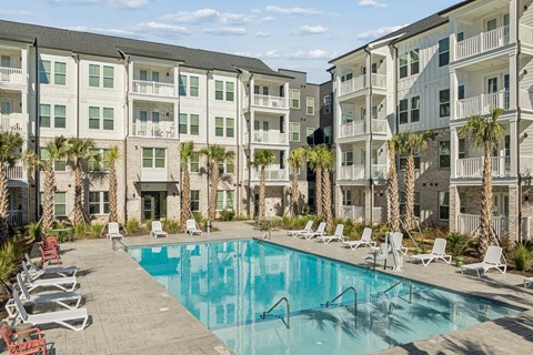 A swimming pool surrounded by lounge chairs and palm trees in front of apartment buildings.