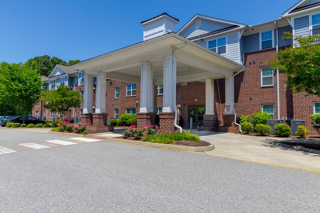 Grand Entrance with Brick Detailing and Garden Beds Spacious front entrance highlighted by tall white columns, brick detailing, and flower-filled garden beds at Snowden Senior Apartments.