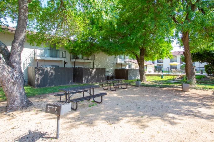 A picnic table is in the middle of a sandy area with trees and a building in the background.