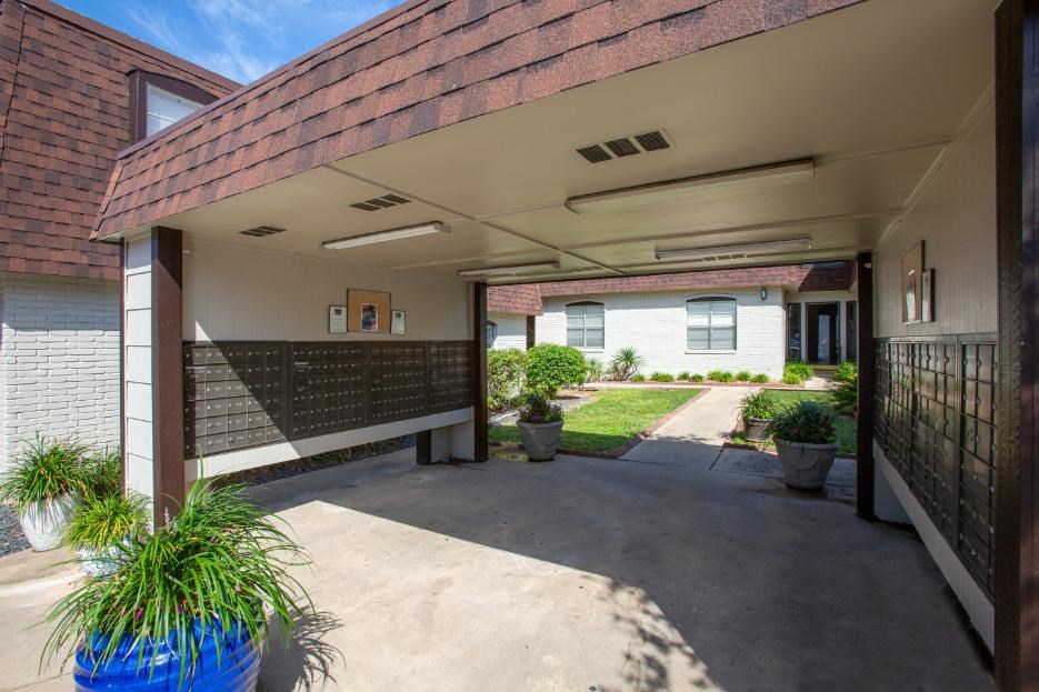 A covered walkway leads to a white building with a brick wall and a black fence.