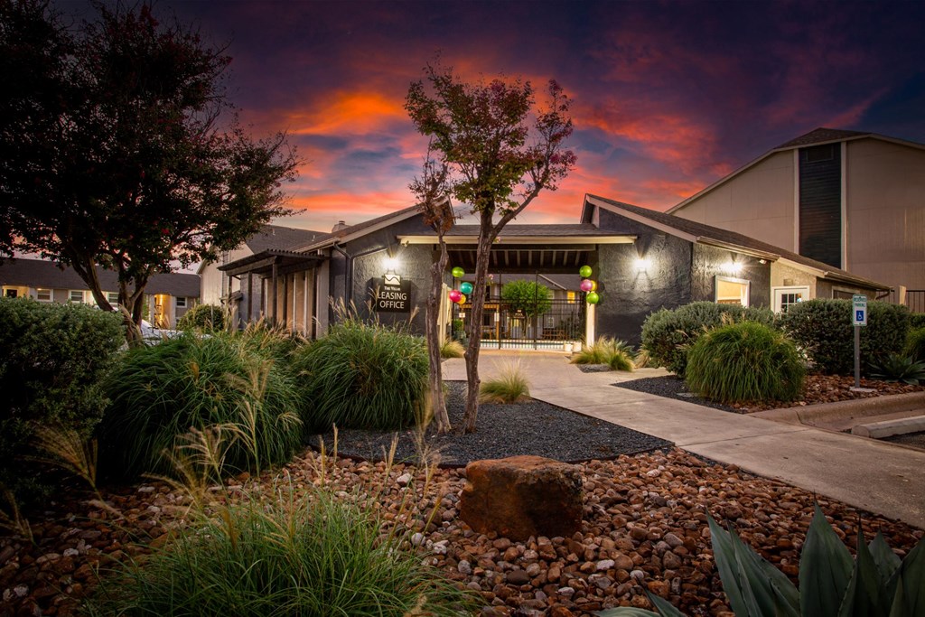 A building with a dark facade is surrounded by a garden with a large rock in the foreground.