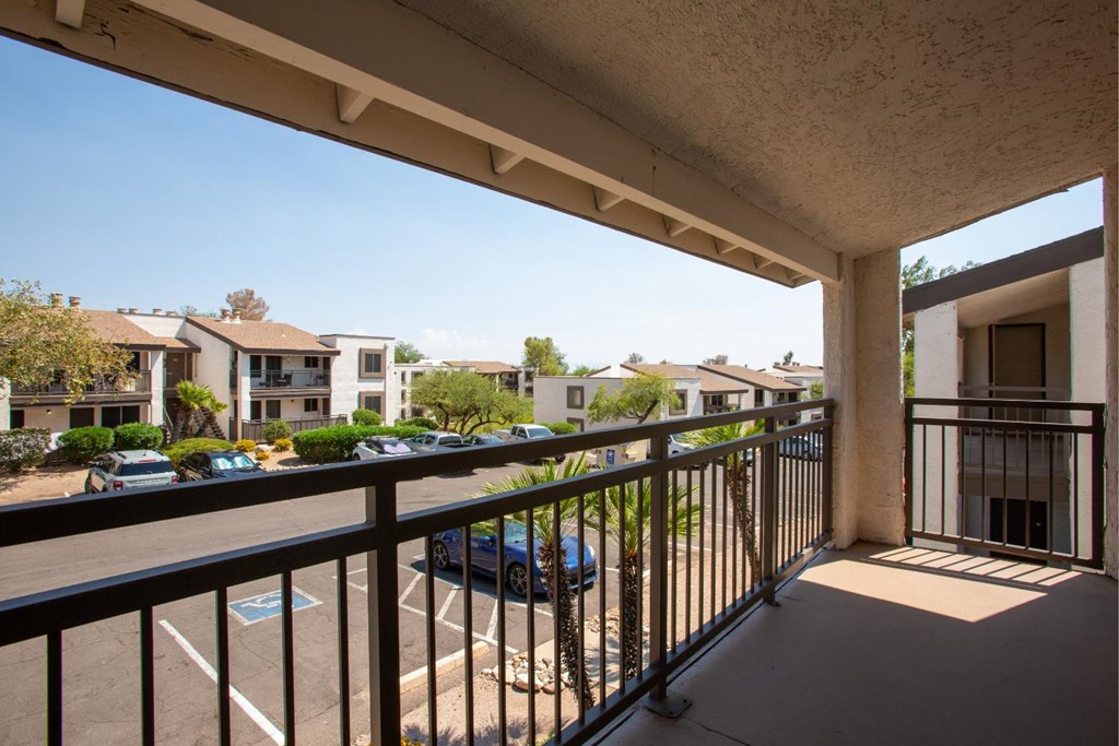 A balcony with a black railing overlooks a parking lot and buildings.