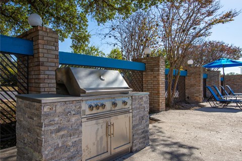 A stone BBQ grill with a blue awning and chairs in the background.