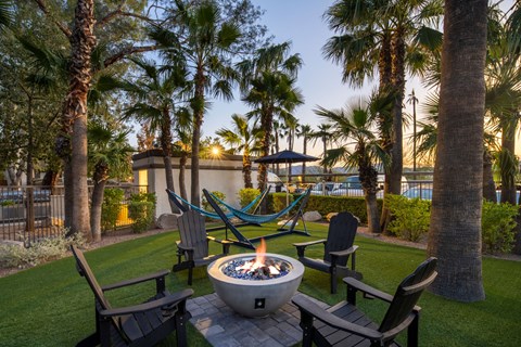A fire pit sits in the middle of a grassy area surrounded by chairs and palm trees.
