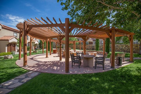 A wooden pergola with a table and chairs is surrounded by a grassy area.
