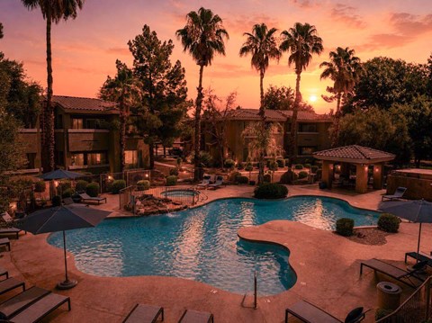 A sunset view of a pool area with palm trees and a building in the background.