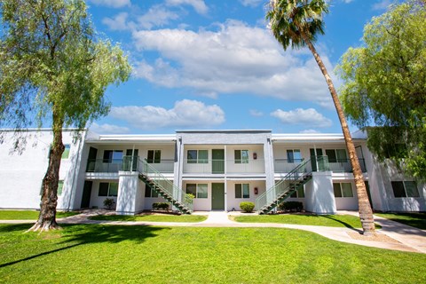 A white building with a balcony on the second floor is surrounded by green grass and trees.