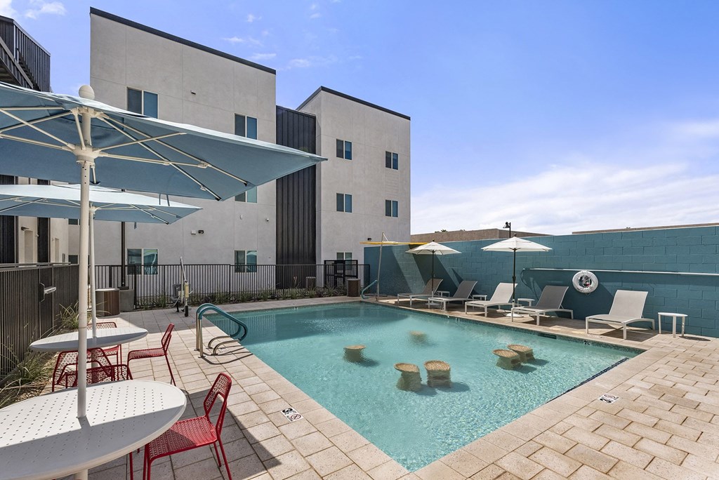 a swimming pool with chairs and umbrellas and a building in the background a swimming pool with chairs and umbrellas and a building in the background at SECOND AVE COMMONS | BRAND-NEW, Mesa, Arizona