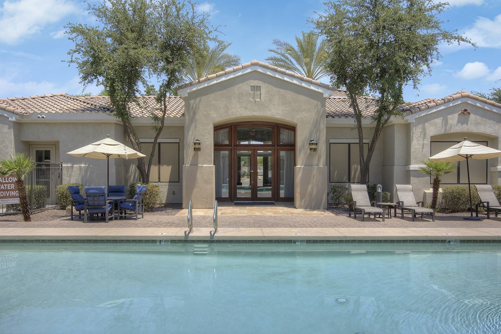 A pool in front of a house with a patio and chairs.