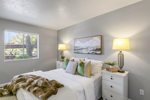 A bedroom with a white bed, a white nightstand with a lamp and a painting on the wall.