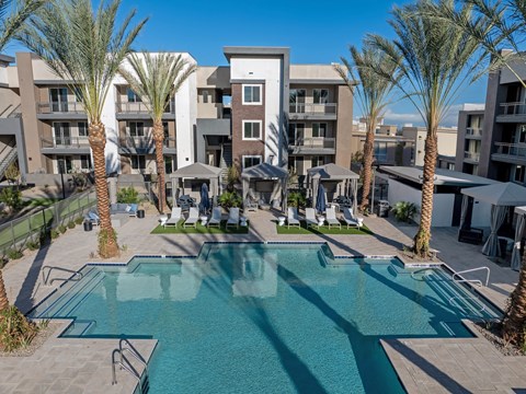 A swimming pool surrounded by palm trees and lounge chairs in front of a multi-story apartment building.