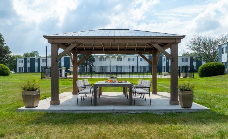 A wooden gazebo with a table and chairs is set up in a grassy area.