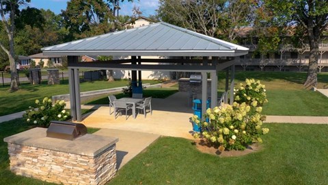 A gazebo with a stone pillar and a fireplace is surrounded by a lawn and trees.