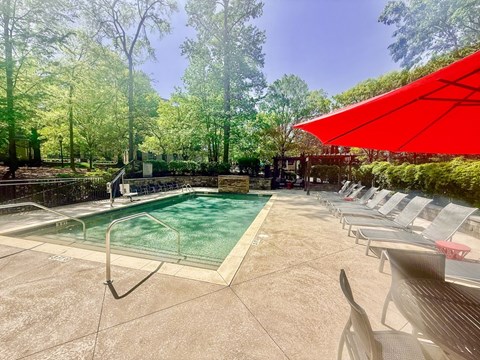 A pool surrounded by trees and chairs under a red umbrella.