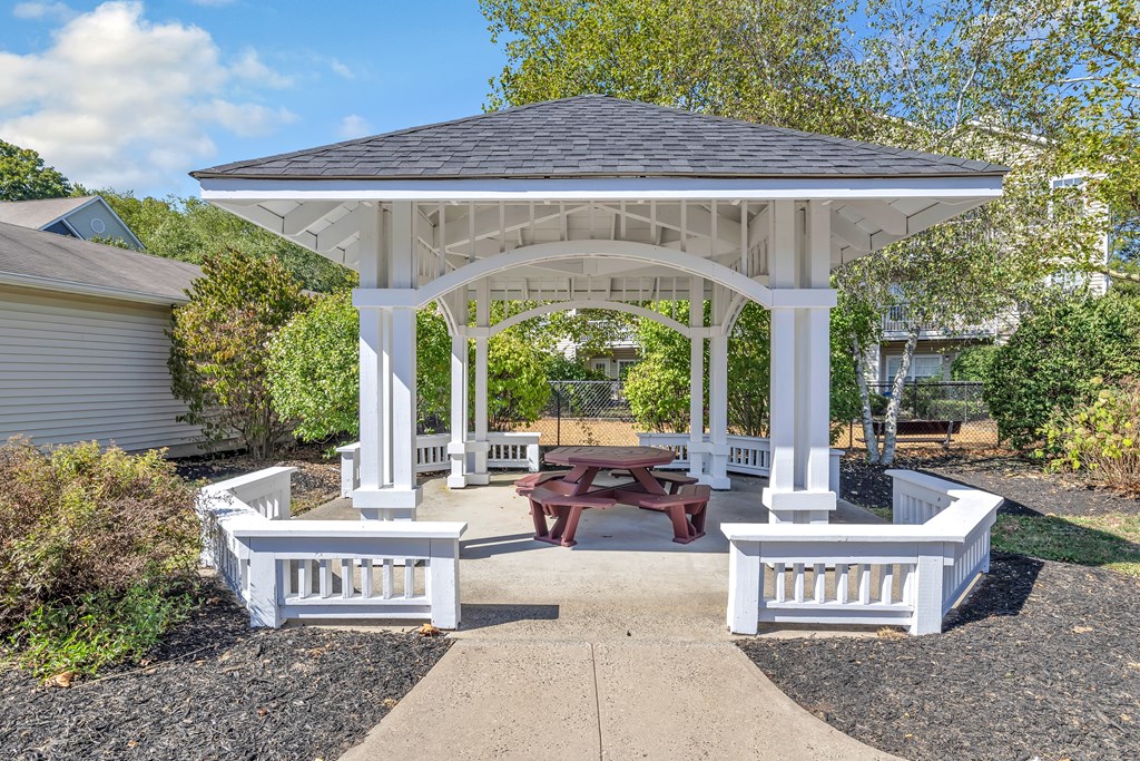 A white gazebo with a bench in front is surrounded by trees.