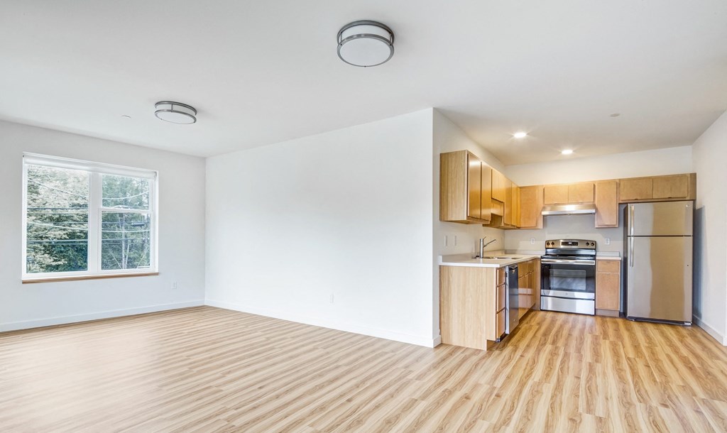 an empty living room with wood floors and a kitchen with stainless steel appliances