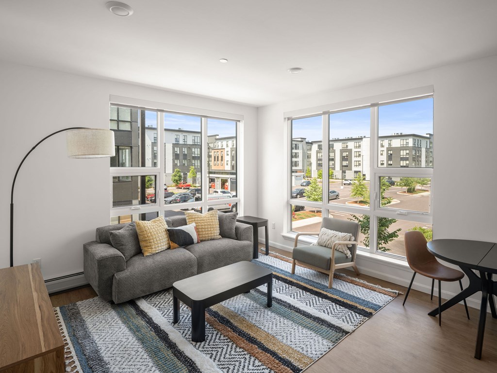 A living room with a grey couch, a coffee table, a chair, and a large window with a view of the street.