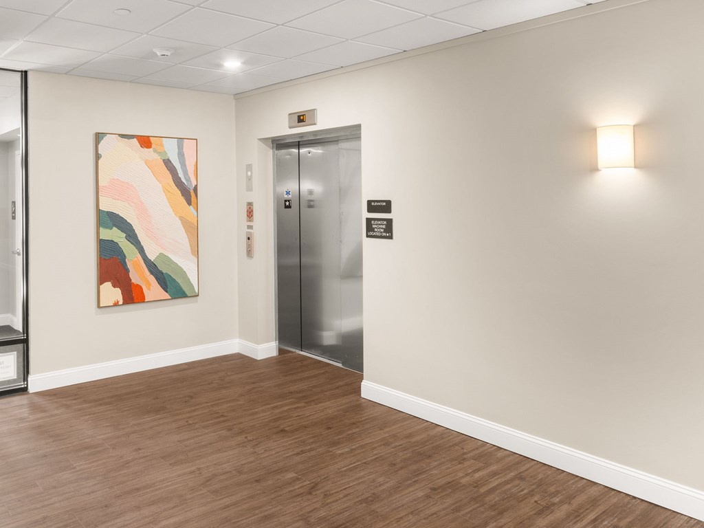 A hallway with a wooden floor, a framed painting on the wall, and a silver elevator.