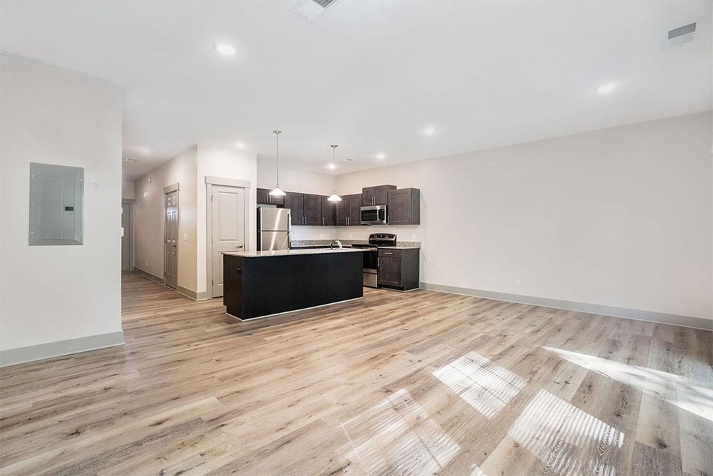 A spacious room with wooden flooring and a kitchen area in the background.