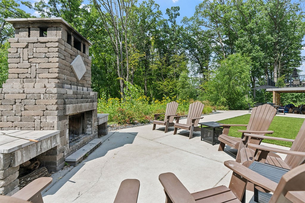 A stone fireplace is in the middle of a patio with wooden chairs around it.