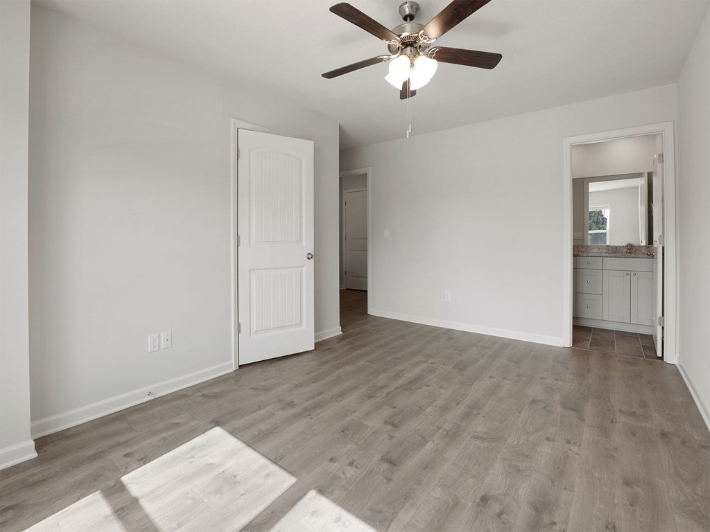 A room with a ceiling fan and light fixture, wooden flooring, and white walls with a door and a mirror.
