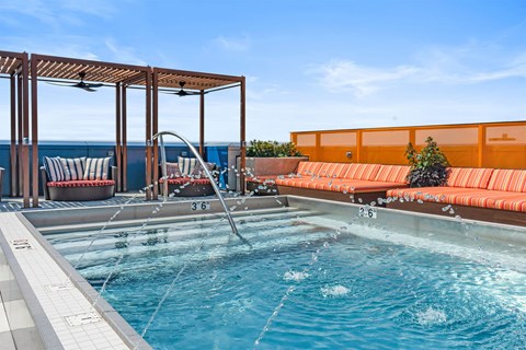 A pool with a waterfall and a bench with orange cushions.