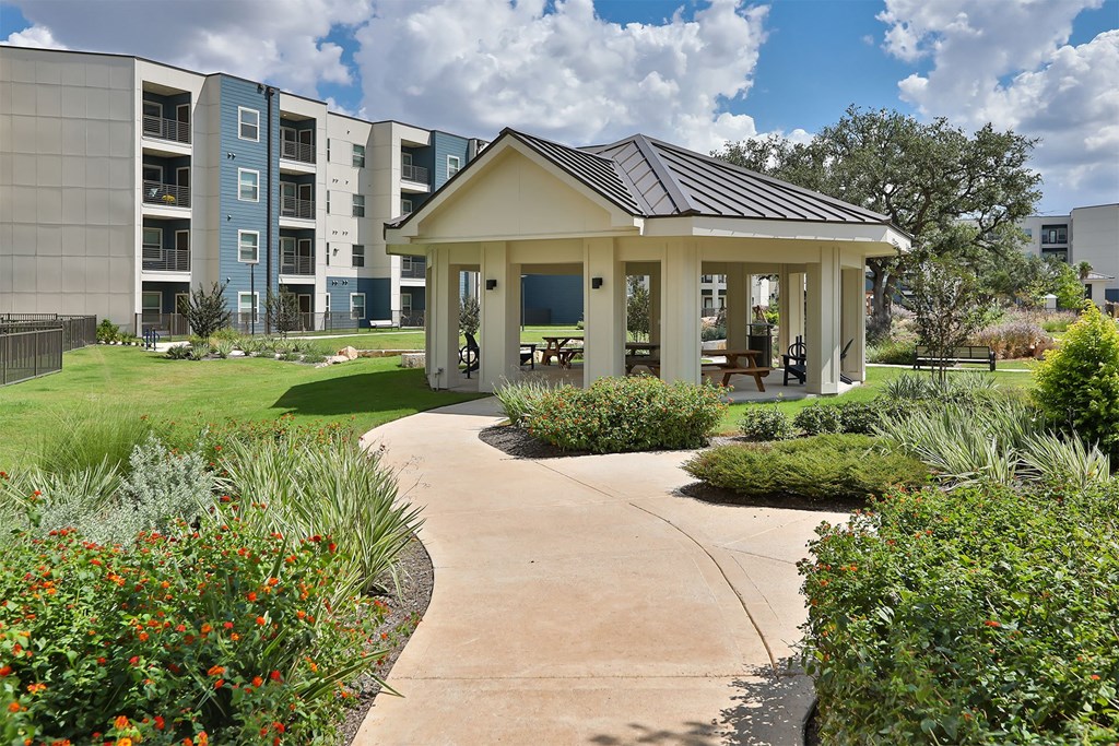 A small pavilion with a roof is surrounded by a concrete walkway and a grassy area with flowers.