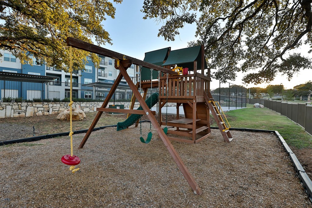 A wooden swing set with a red swing sits in a gravel backyard.
