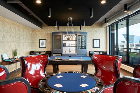 A poker table surrounded by red chairs in a room with a pool table and a view of the city.