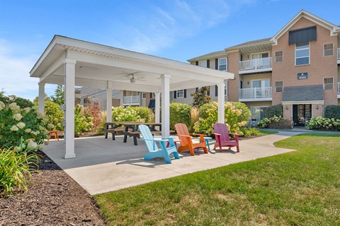 A white pergola is in front of a building with a patio and chairs.