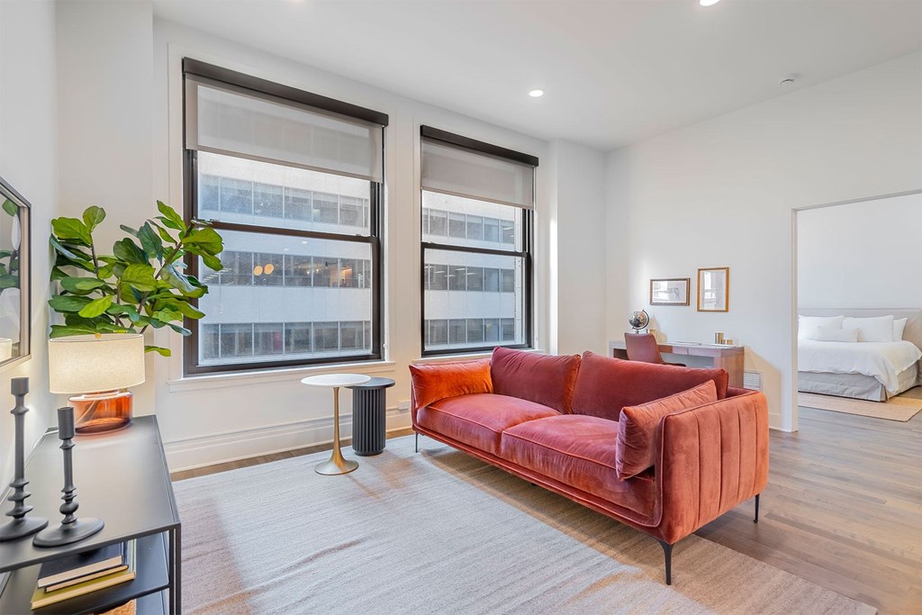 A red couch is in the middle of a white room with a large window.