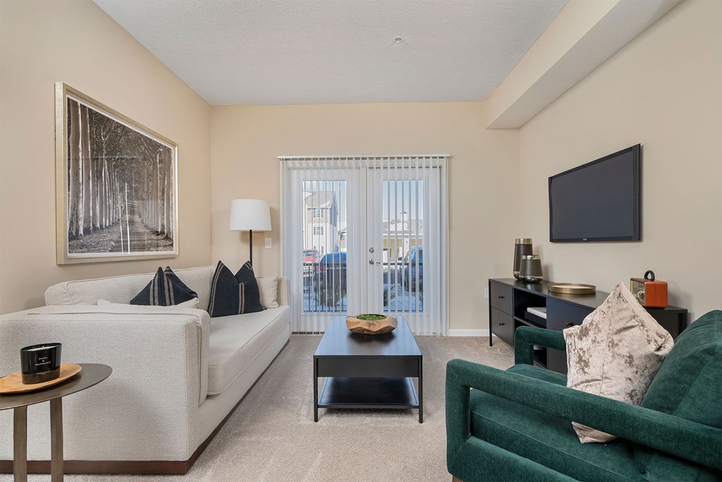 A living room with a white couch, a green couch, a coffee table, a side table, a lamp, a painting, a flat screen TV, and a sliding glass door.